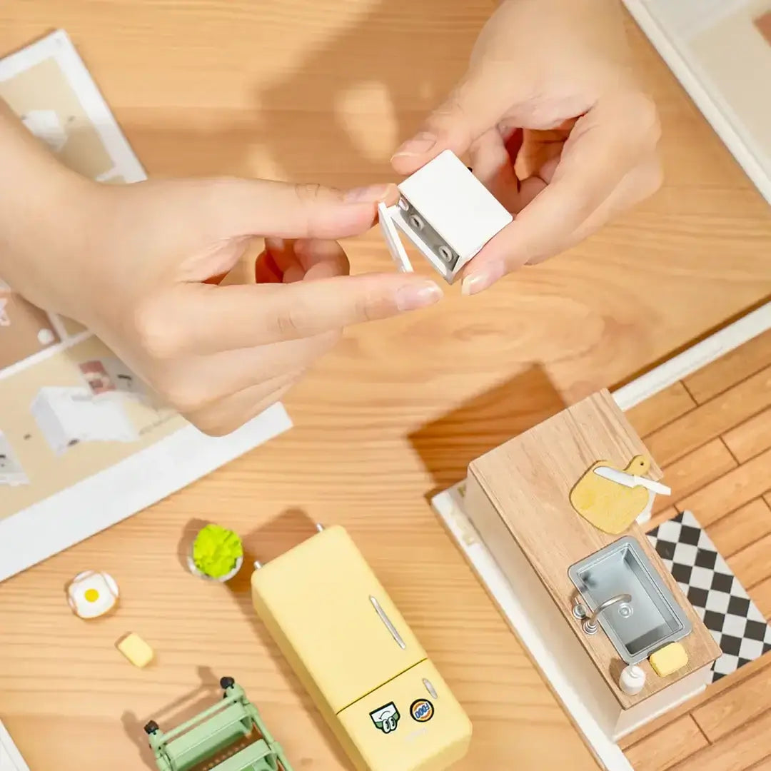 Hands assembling a miniature kitchen appliance from the Happy Meals Kitchen DIY Plastic Miniature House kit with other miniature items around.